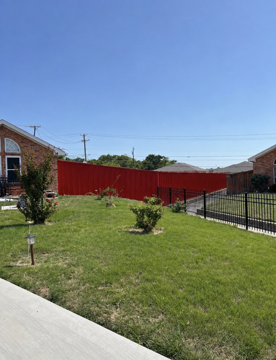 Bright red corrugated metal privacy fence with a black metal fence, green lawn, and brick houses under a blue sky in Killeen TX by Martinez Construction fencing contractor